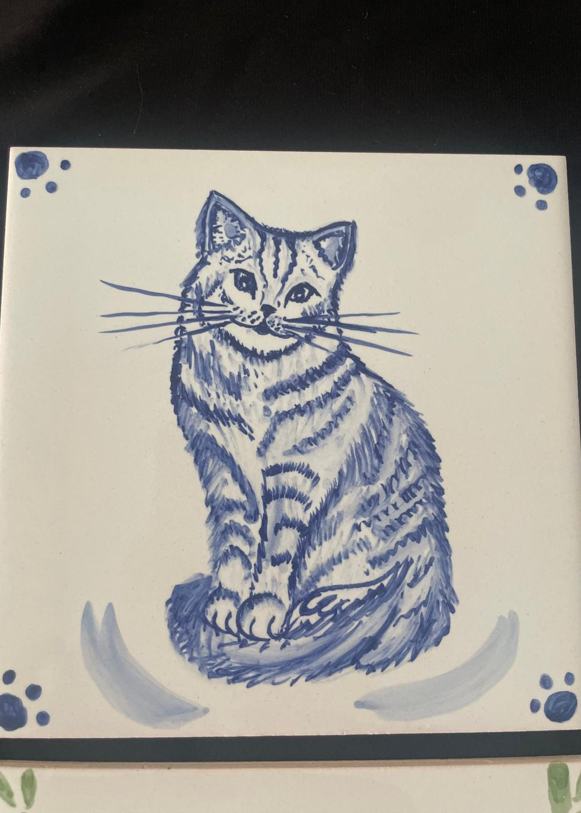 A square ceramic tile featuring a blue hand-painted illustration of a sitting tabby cat, with blue paw print details in the corners on an off-white background.