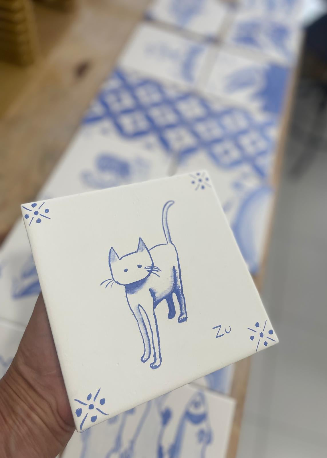 A hand holds a square white ceramic tile featuring a blue line drawing of a cat and decorative corner motifs. More matching blue-and-white patterned tiles are blurred in the background on a wooden surface.
