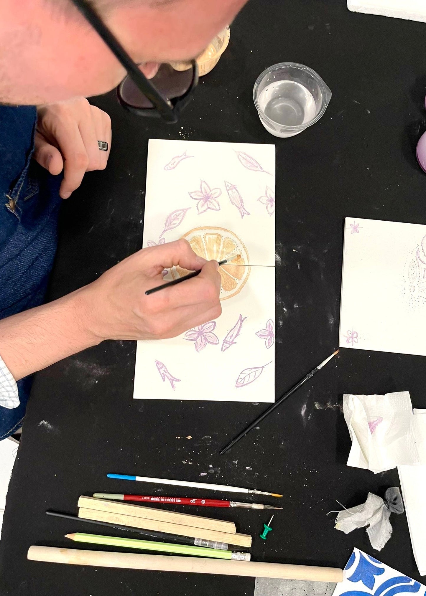 Person drawing on a piece of paper with various art supplies on a black surface