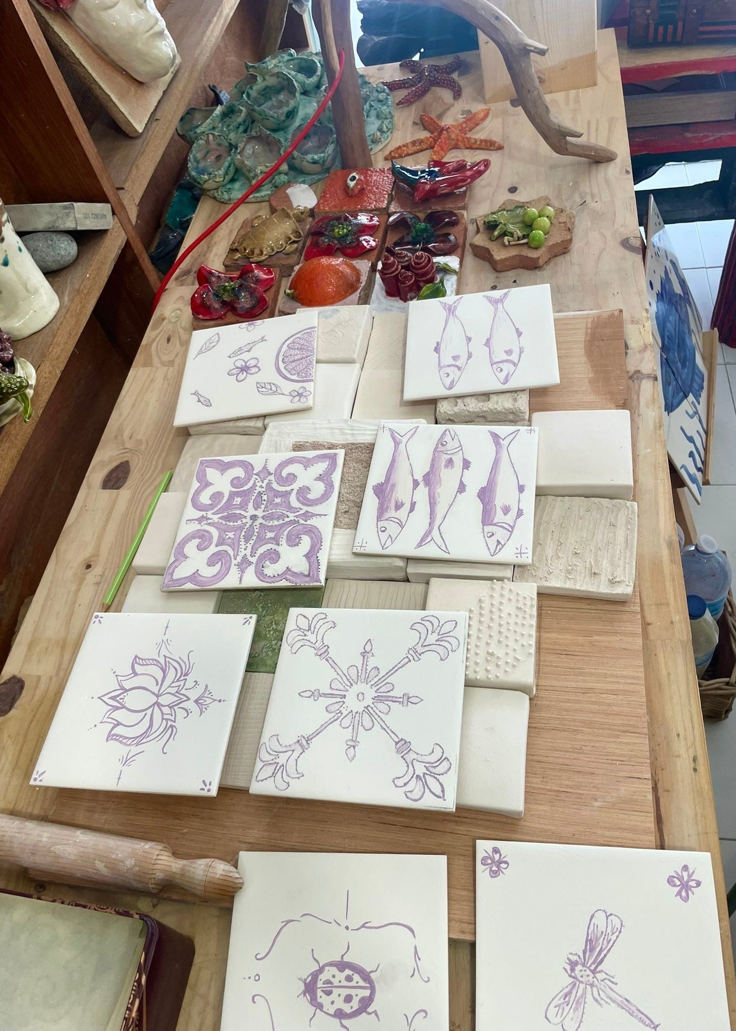 A display of hand-painted ceramic tiles with purple designs including fish, lotus, snowflake, dragonfly, and abstract patterns, arranged on a wooden table.