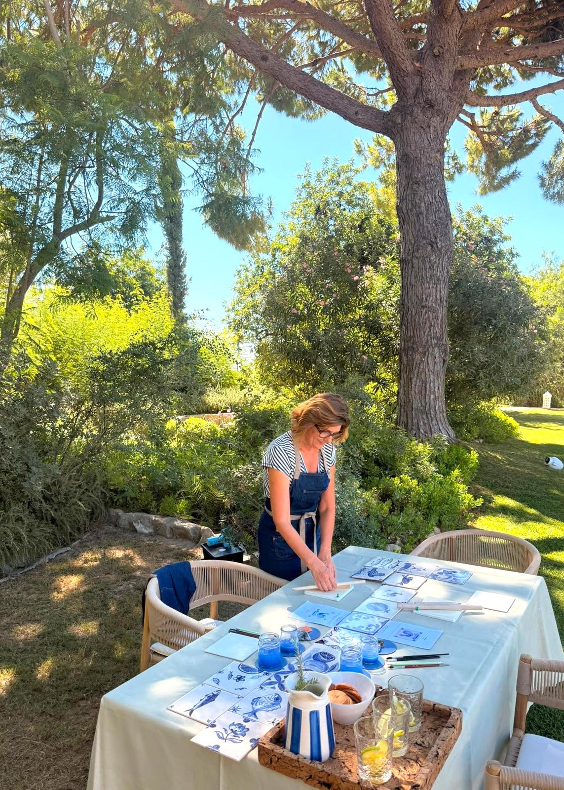 A woman in a blue apron stands at an outdoor table covered with blue-and-white patterned sheets, glasses, and bottles, set in a sunny garden with trees and greenery in the background.