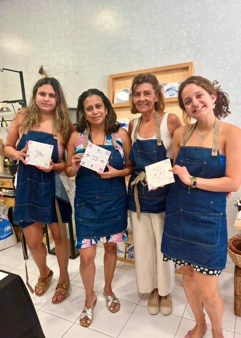 Four women standing together in a pottery studio, each wearing a blue denim apron and holding small square ceramic pieces. The background shows pottery tools, shelves, and a tiled floor.