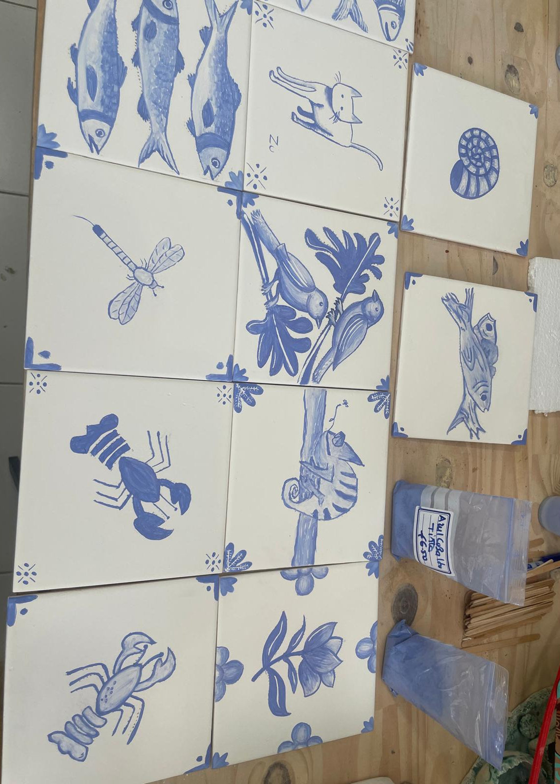 A set of square ceramic tiles with blue illustrations of marine life and nature motifs on a white background, arranged on a wooden surface next to a plastic bag labeled 'Pistachio'.