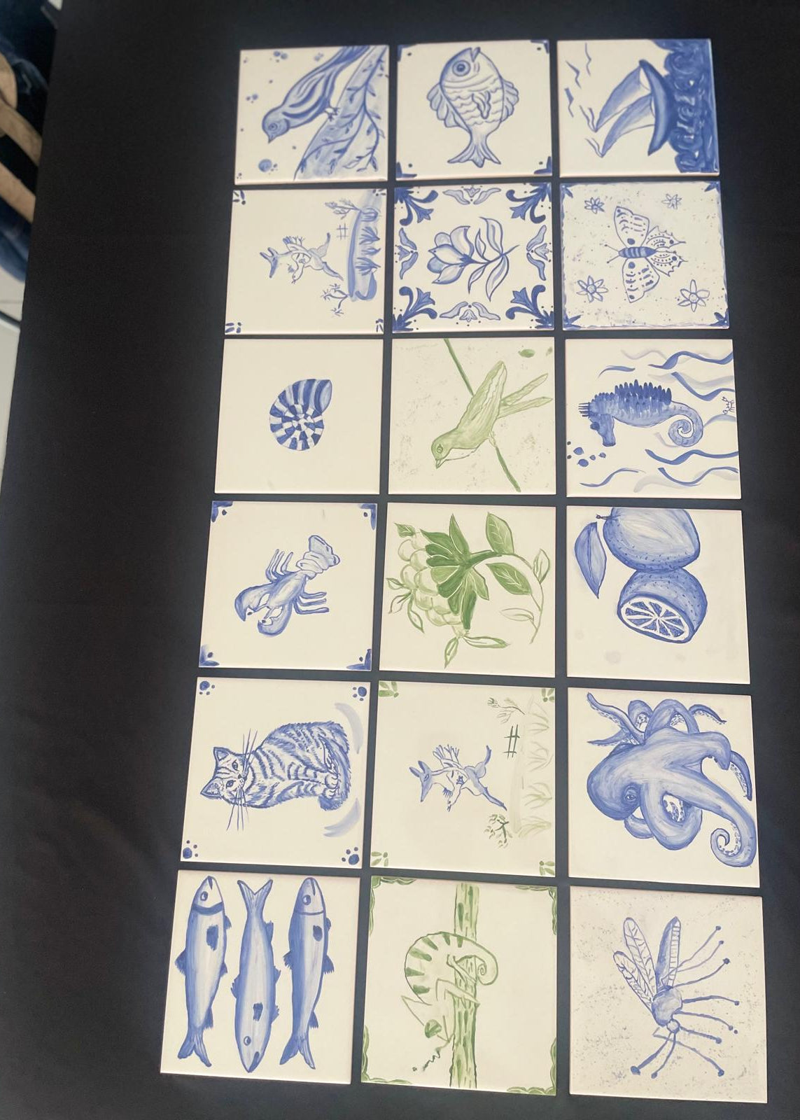 A set of 15 square ceramic tiles arranged in a 3x5 grid on a black surface. Each tile features a blue and green hand-drawn illustration of marine life, plants, and sea creatures on an off-white background.