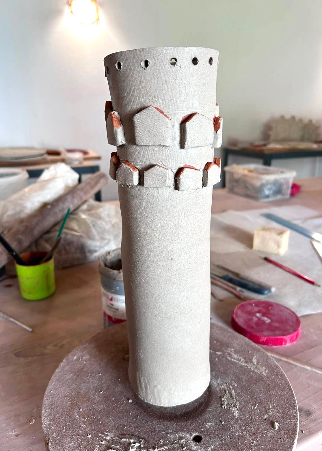 A handmade ceramic cylinder with a textured, matte finish and decorative relief patterns near the top, displayed on a pottery wheel in a workshop setting.