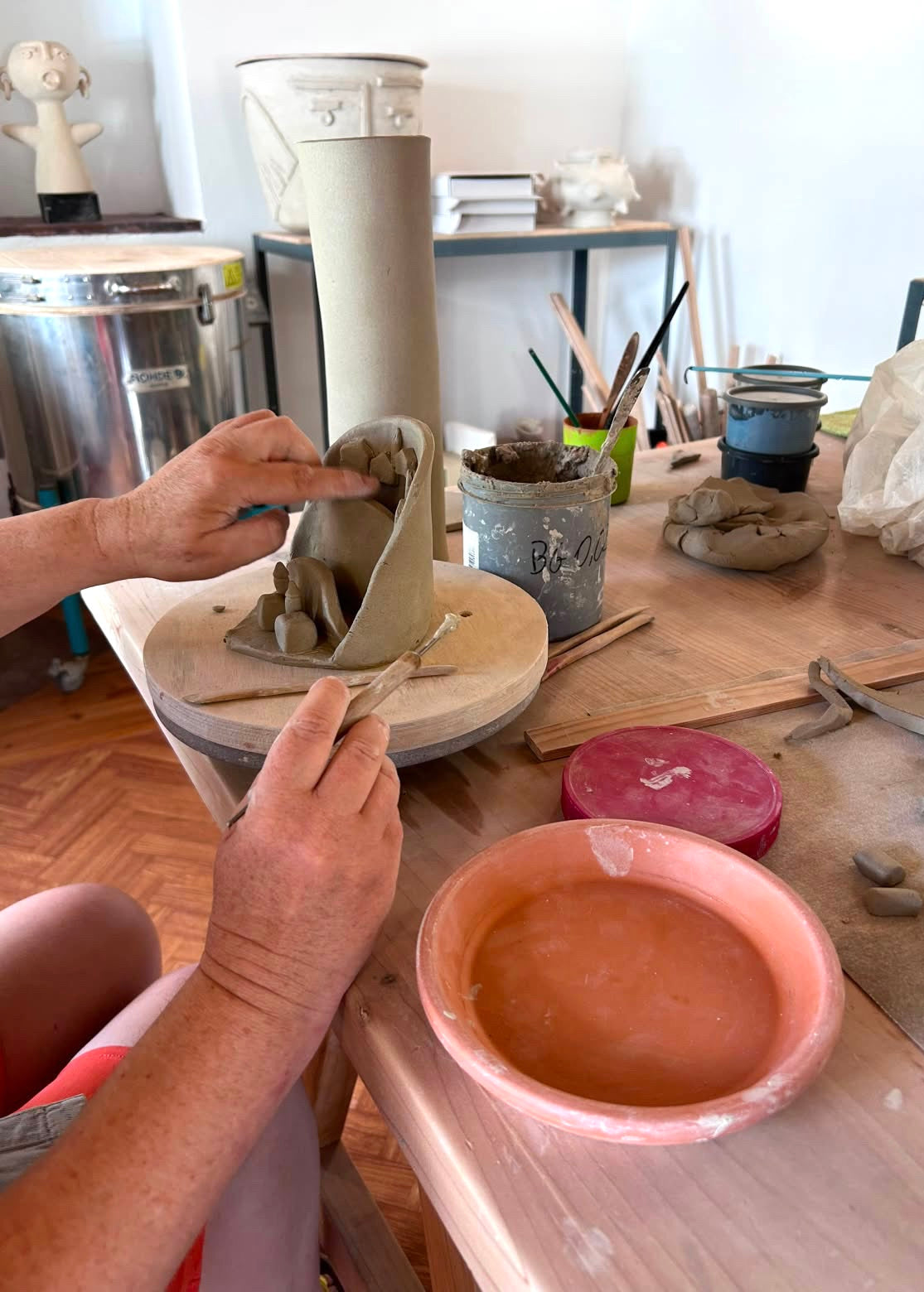 A person's hands sculpting a clay vessel on a pottery wheel, with various clay tools, a pink clay bowl, and other pottery items visible in a workshop setting.