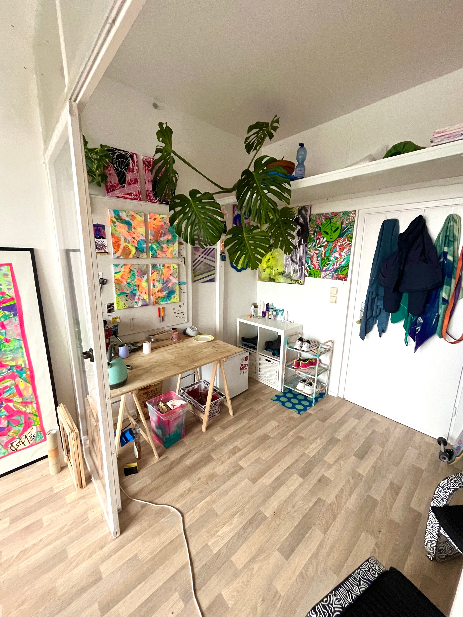 Small home office with a desk, shelves, and colorful artwork on a wooden floor.