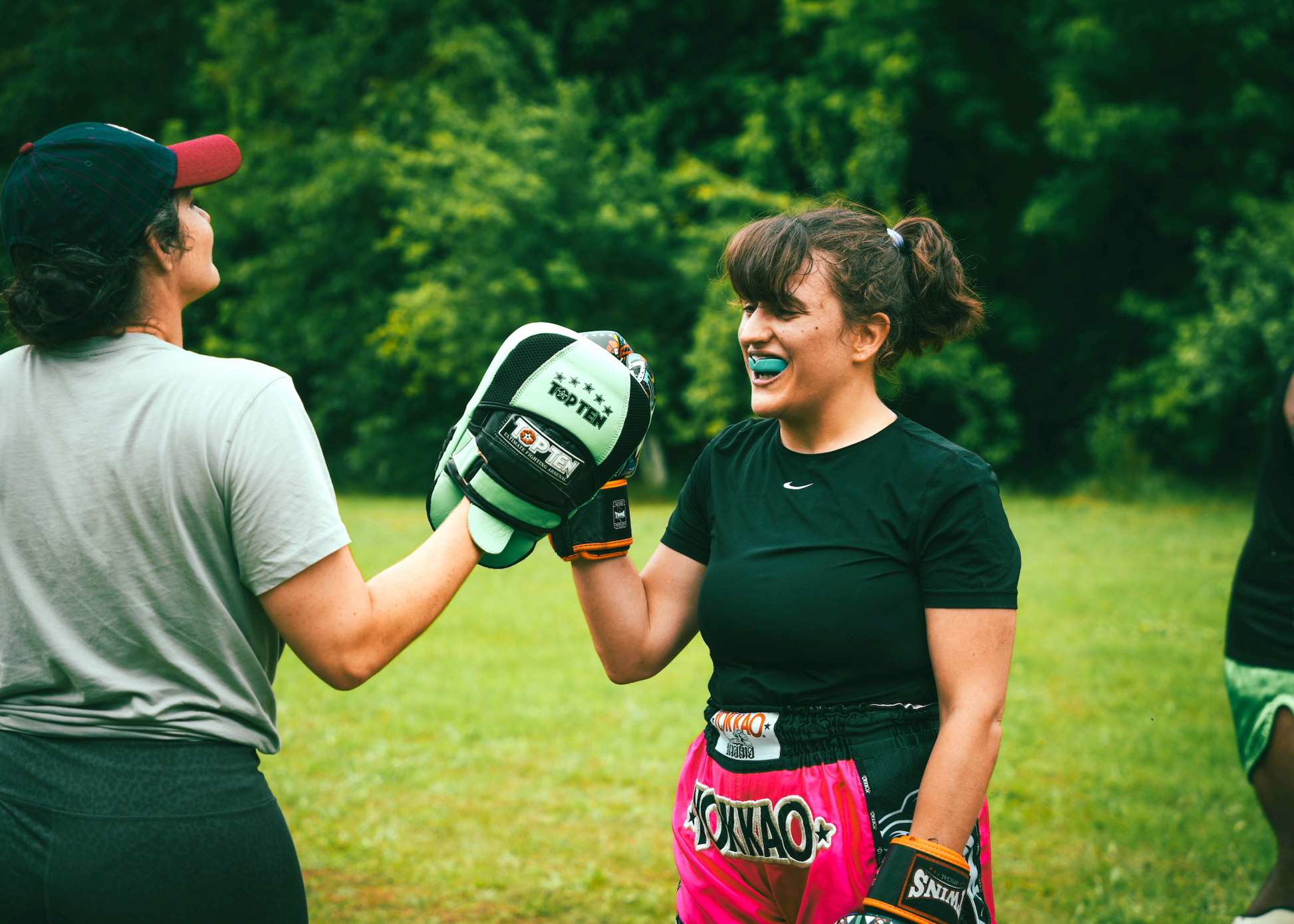 Two people in a grassy outdoor setting, one wearing boxing gloves and the other in a gray shirt.