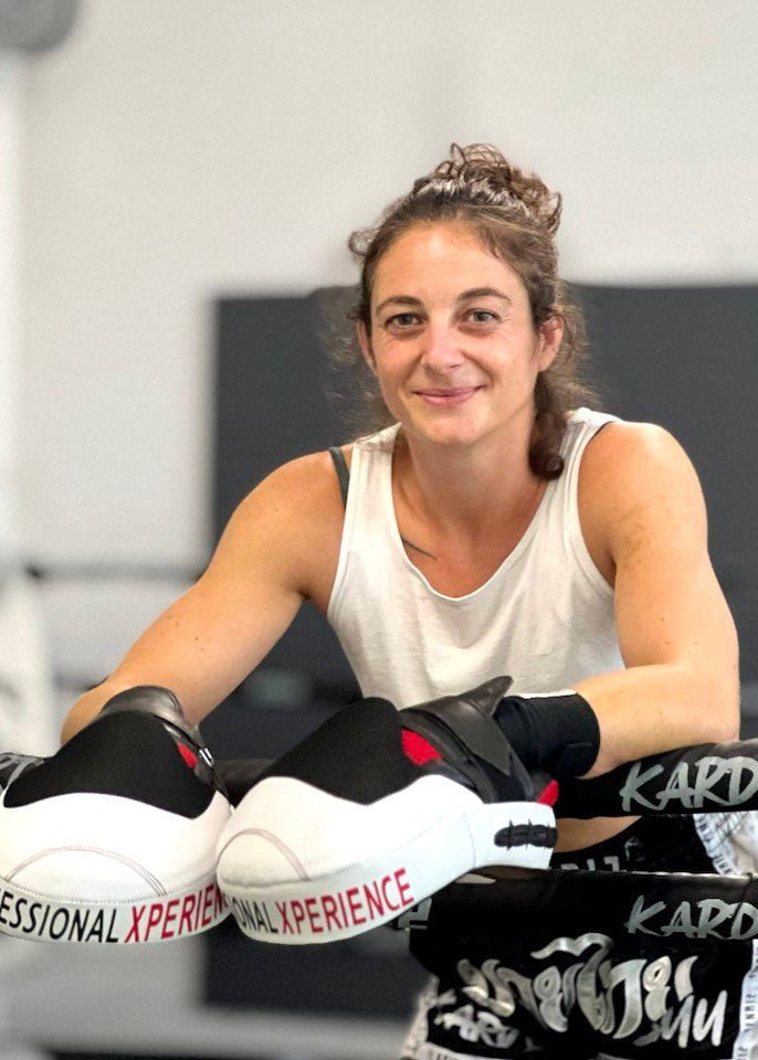 Woman in a white tank top and boxing gloves sitting in a boxing ring.