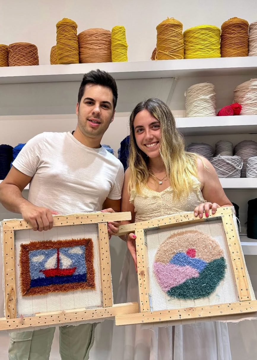 Two people holding framed woven artworks in a yarn store.