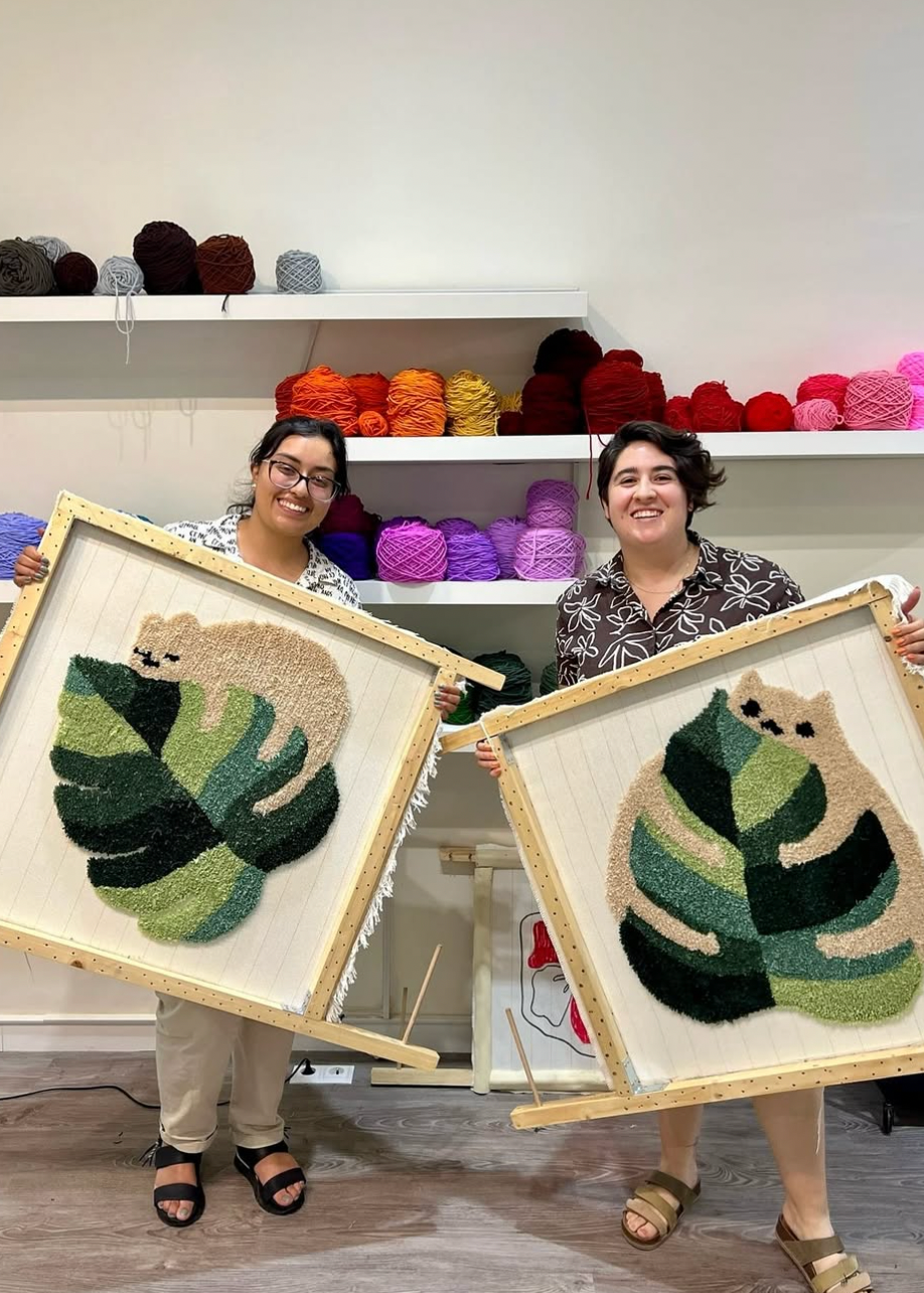 Two people holding framed needlepoint artworks of animals in a yarn store.