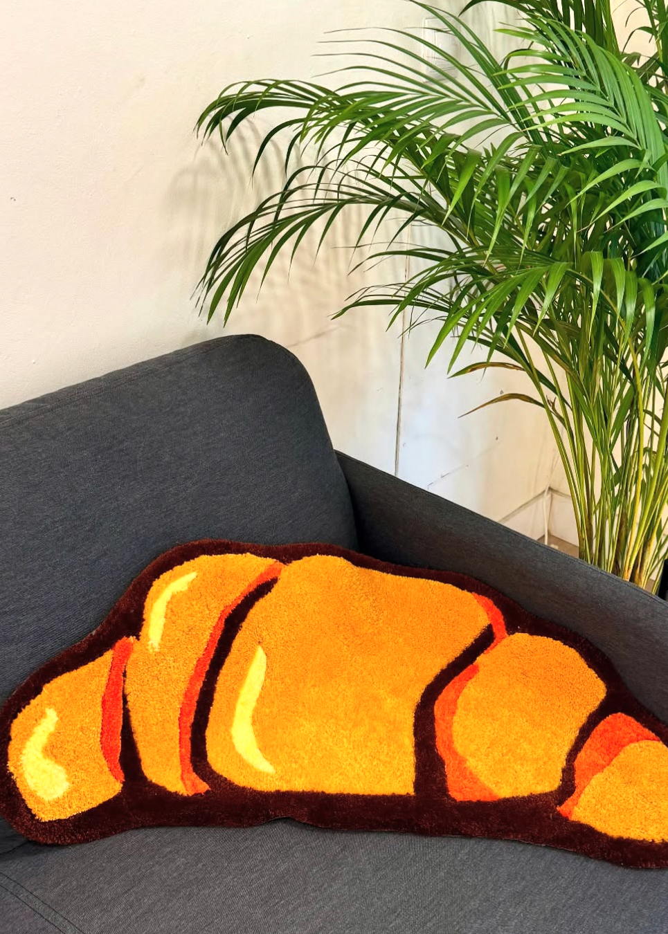 Croissant-shaped cushion on a gray sofa with a plant and a drink in the background