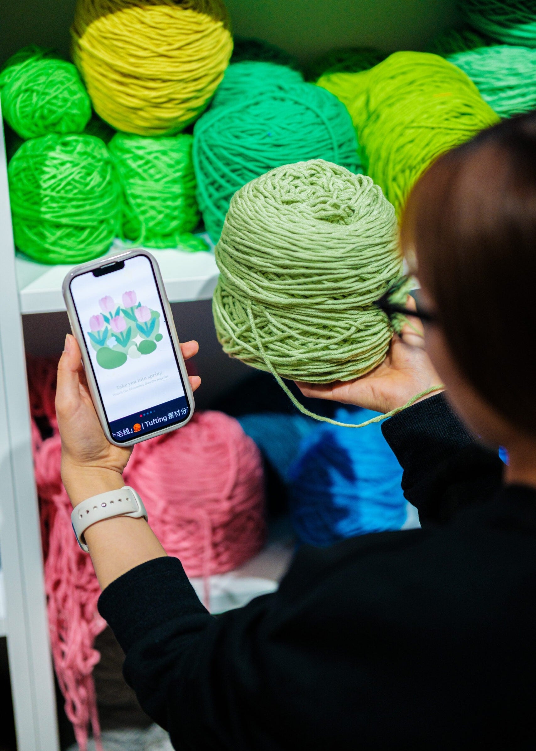 Person holding a skein of green yarn and a phone with colorful yarn balls in the background