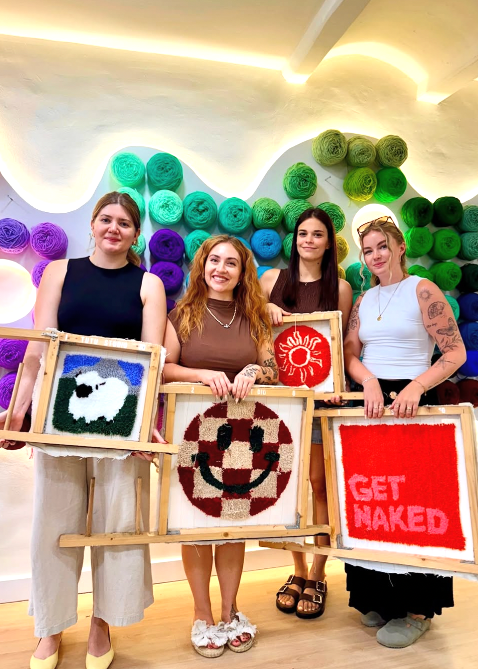 Four women holding framed artworks in front of a colorful yarn wall.