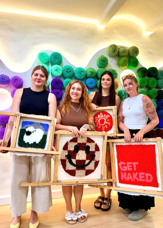 Four women holding framed artworks in front of a colorful yarn wall.