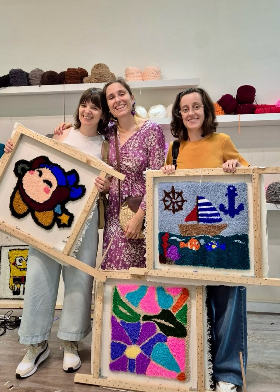 Three women holding up framed needlepoint artworks in a store setting.