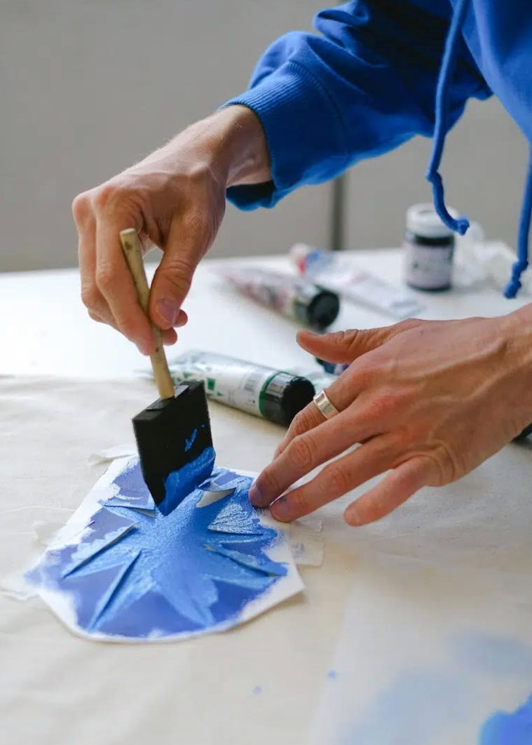 A close-up of a person's hands using a paint brush to apply blue paint onto a white surface, with paint tubes and a small jar in the background.