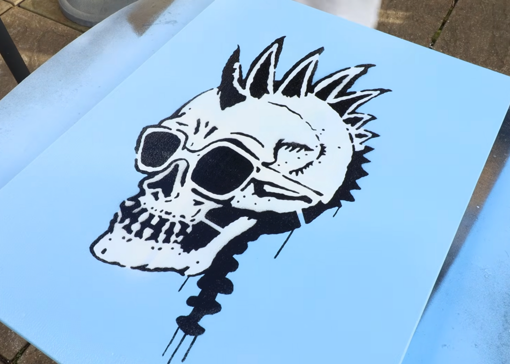 A black and white stencil or decal of a skull with a mohawk and sunglasses, shown on a light blue sheet of vinyl or metal over a tiled floor.