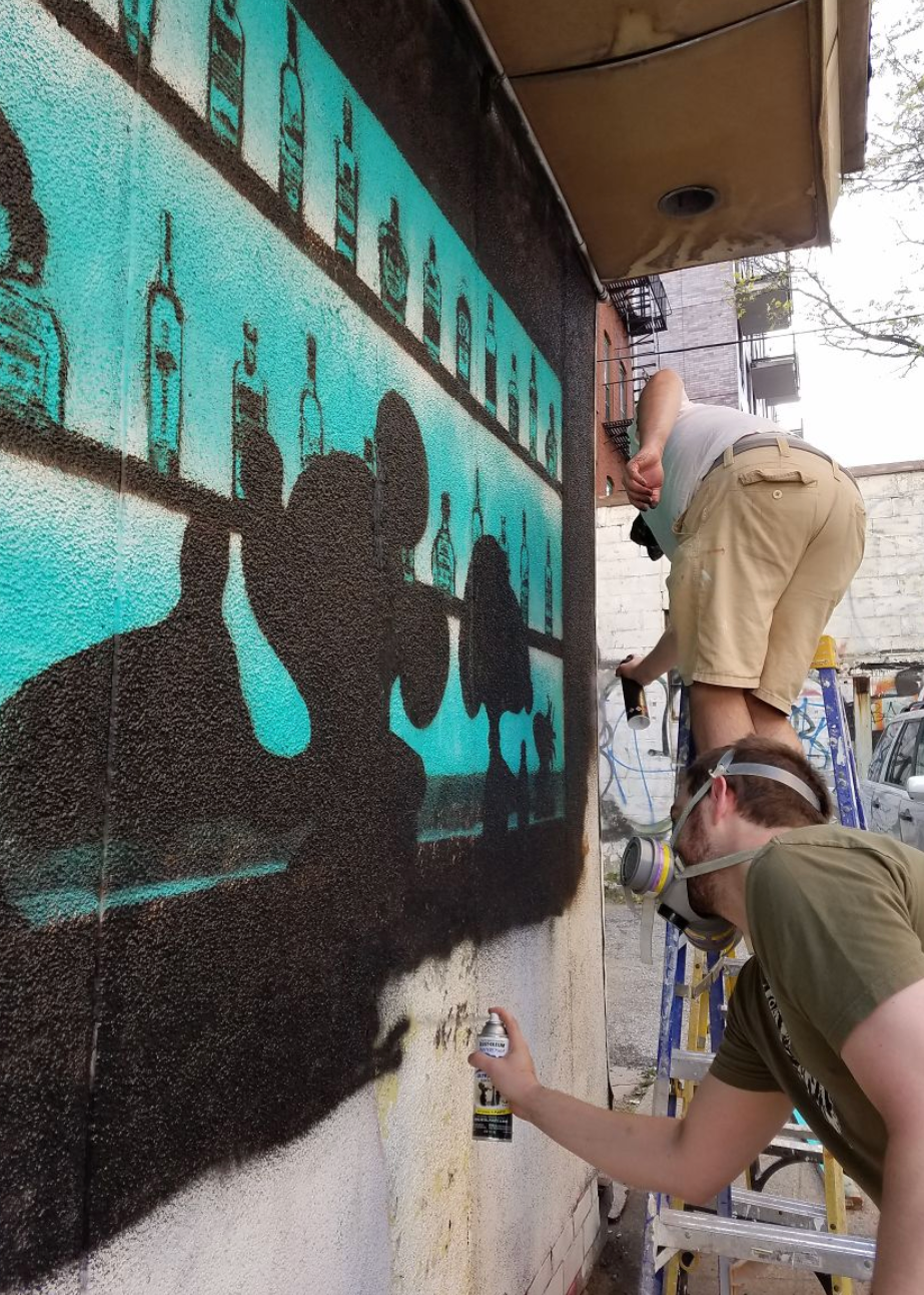 Two people spray painting a mural on an outdoor wall, with one person standing on a ladder and the other holding a spray can in the foreground. The mural features black silhouettes of figures and bar bottles against a turquoise background.