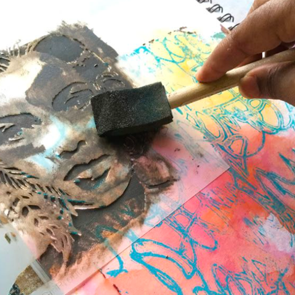 A close-up of a hand using a black foam stamping block to transfer a dark charcoal portrait onto a colorful paper sheet with blue, pink, and orange scribbled artwork.