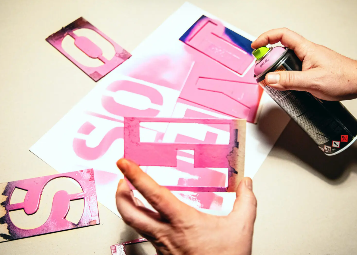 Hands holding a black spray paint can and stencil pieces over a white sheet, creating a pink graffiti-style letter design on a beige surface.
