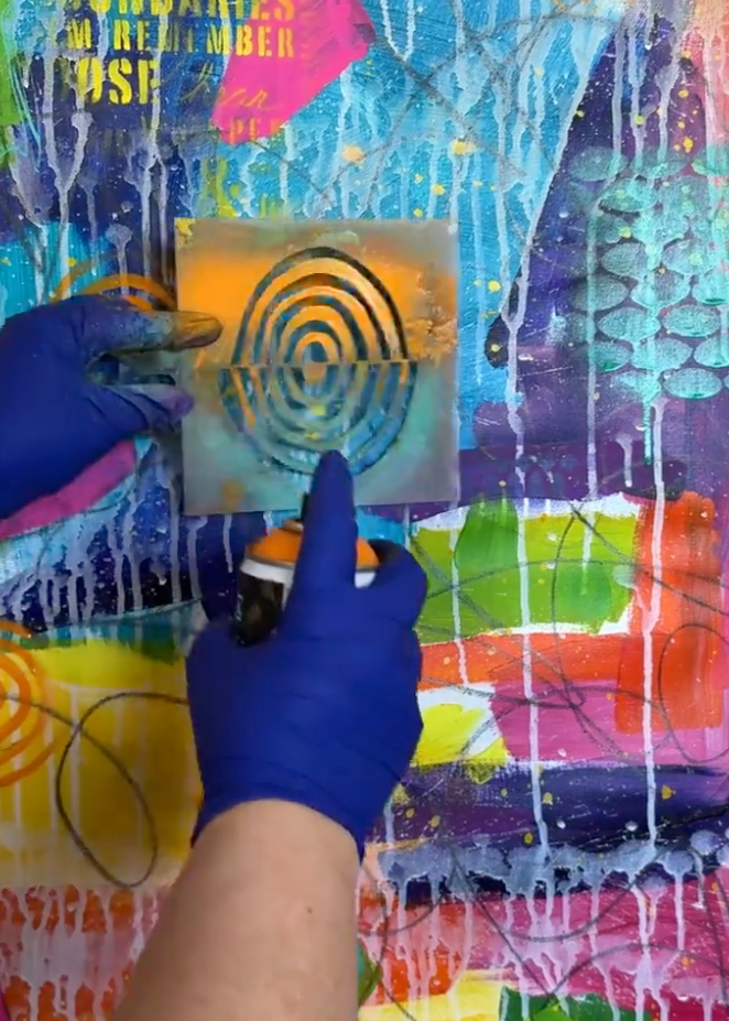 A person wearing blue gloves holds a stencil with a spiral pattern over a colorful abstract paint-splattered surface, creating a stamped design.