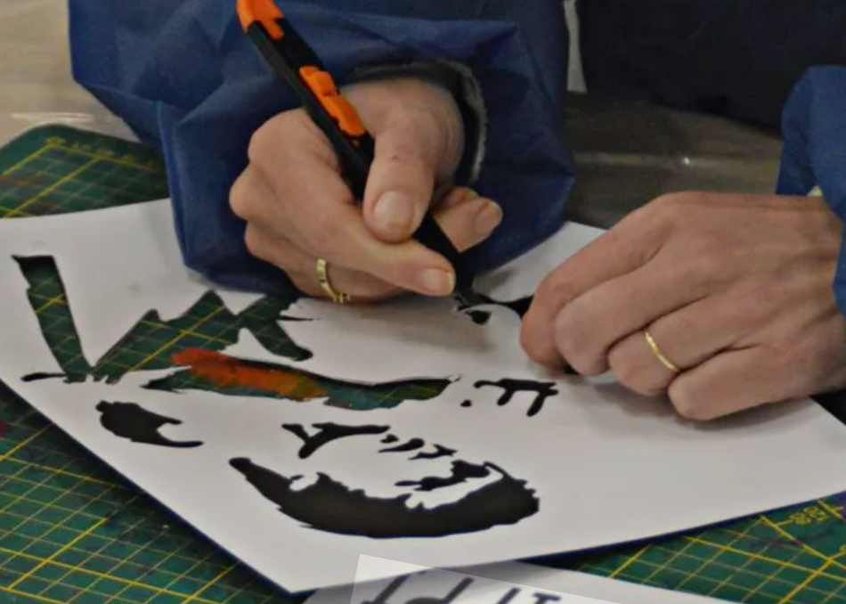 A close-up of a person's hands wearing blue protective sleeves, using a black and orange pen to draw or write on a white sheet of paper over a green cutting mat.