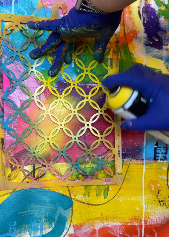 A close-up of hands wearing blue painter's gloves applying yellow spray paint over a colorful, patterned canvas or art print featuring interlocking circular shapes.