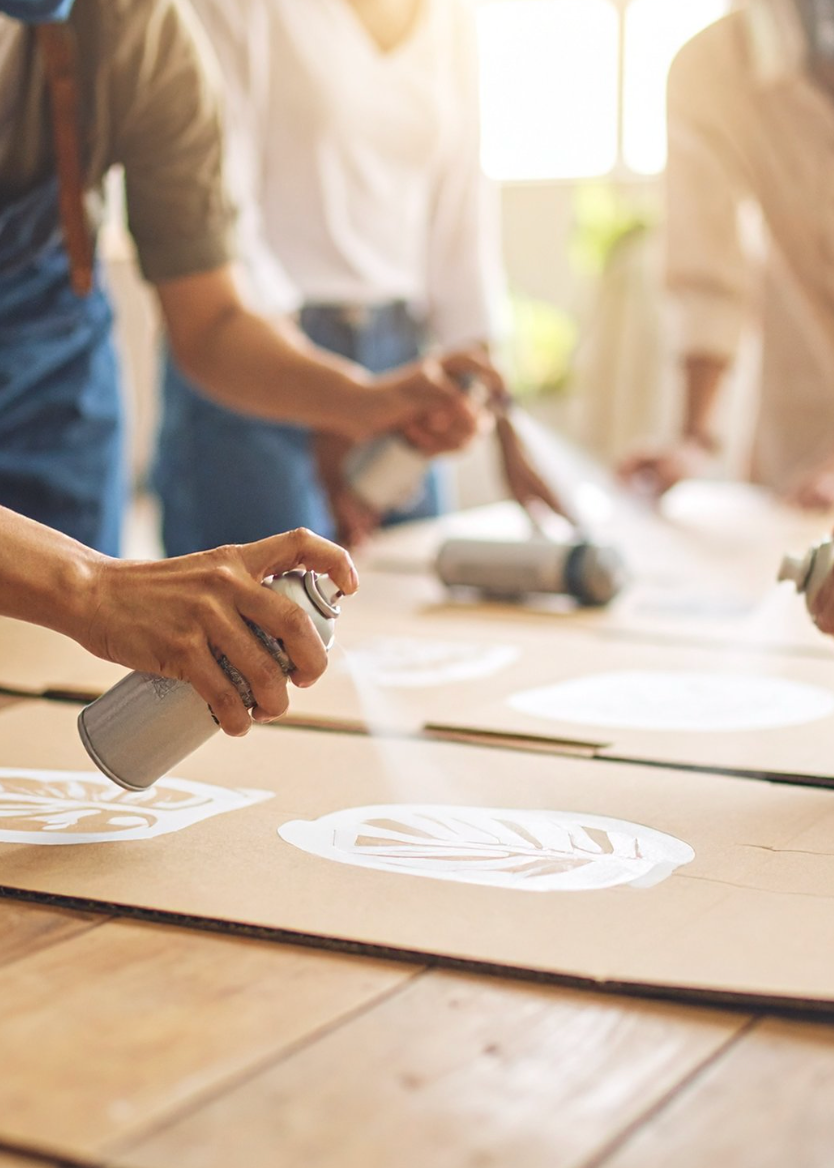 A close-up of a person's hand holding a silver spray paint can and painting a white stencil design onto a large sheet of brown cardboard on a wooden table. In the softly blurred background, other people are visible working with paint and materials in a bright, creative space.