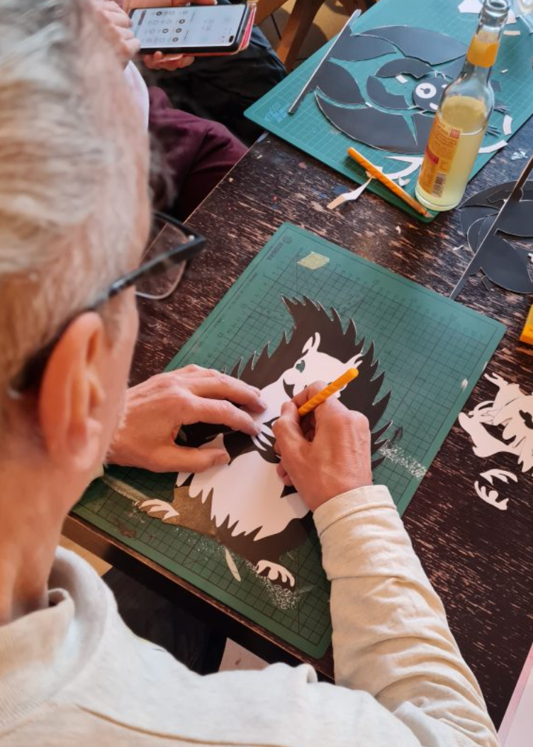 A person wearing glasses and a light beige sweatshirt is carving a detailed animal, likely a hedgehog, from a green mat on a dark wooden table. Bottles and other carving supplies are visible in the background.