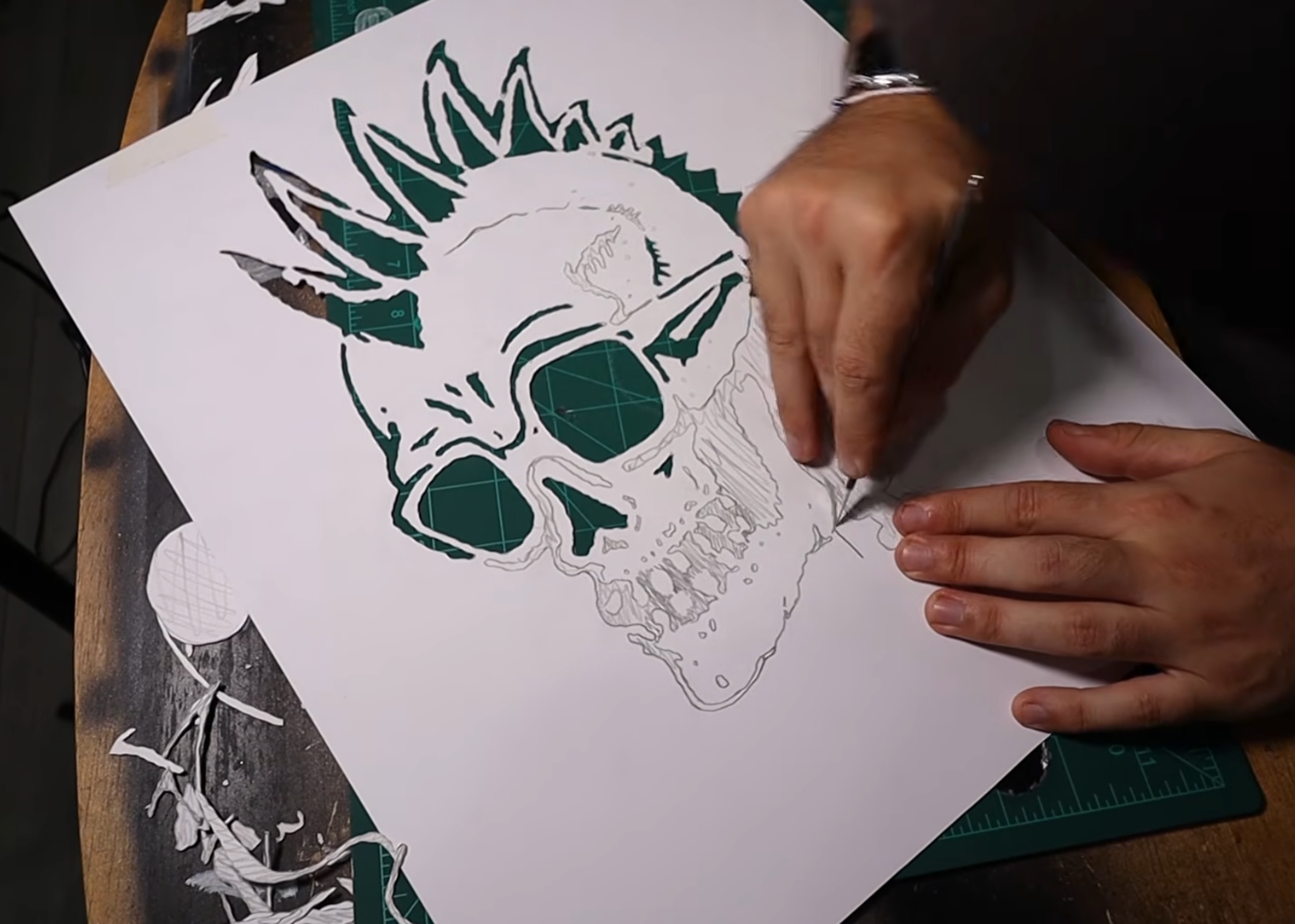 A close-up of a person's hands carving a detailed skull design with a spiked mohawk into a white sheet of paper, using a dark green ink or stamp-like material.