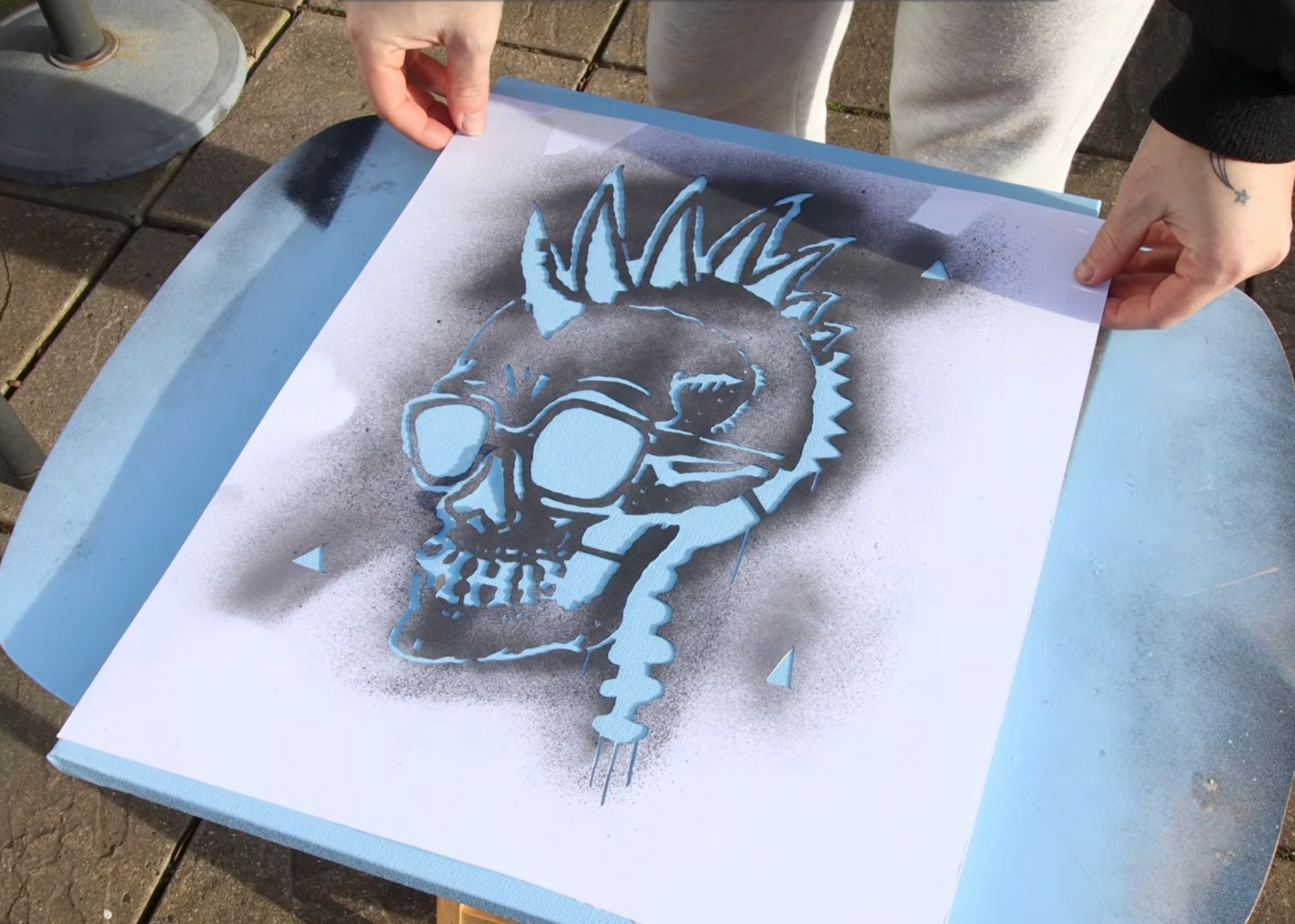 A person holds a sheet of stencil paper over a blue spray table, showing a black and gray airbrushed skull design with a mohawk and aviator sunglasses.