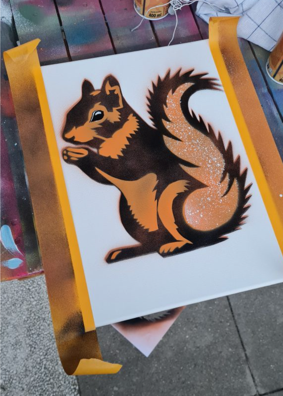 A square white canvas or board featuring a stylized squirrel illustration in black, orange, and glittery gold, photographed outdoors on a painted wooden surface with tape holding the piece in place.