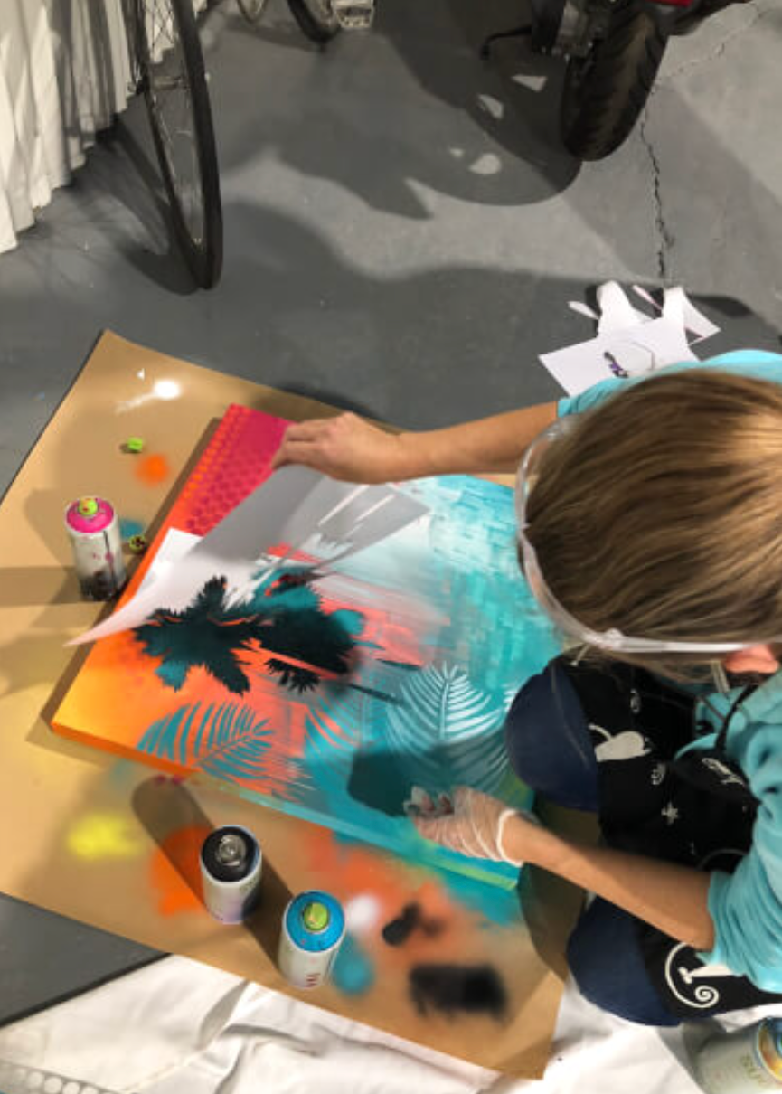 A person wearing a face shield and gloves is painting a tropical scene with palm trees and leaves in turquoise, orange, and black on a canvas laid on a cardboard table. Spray paint cans and other painting supplies are visible on the table, with a workshop or studio floor in the background.