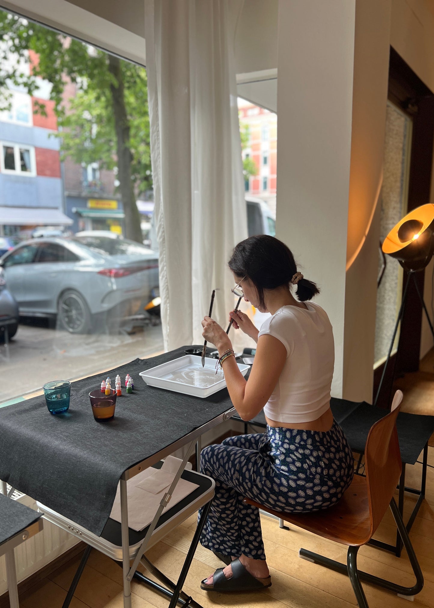 A woman sits at a table by a window, holding a calligraphy brush. She is wearing a white crop top and blue patterned pants.