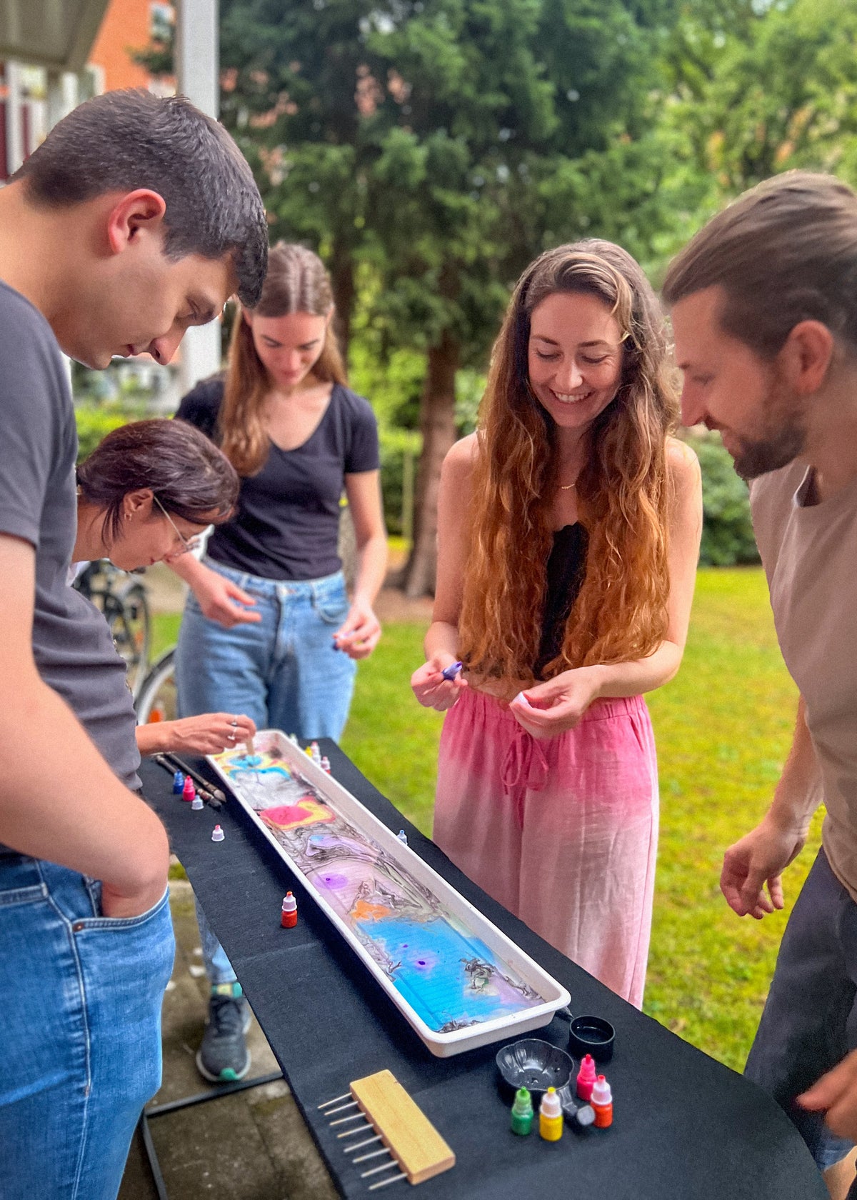 A group of five people gathered outdoors around a long tray filled with colorful powder or paint, appearing to mix or apply it. The scene is set in a grassy backyard with trees in the background, and the people are focused on the tray on a black table.
