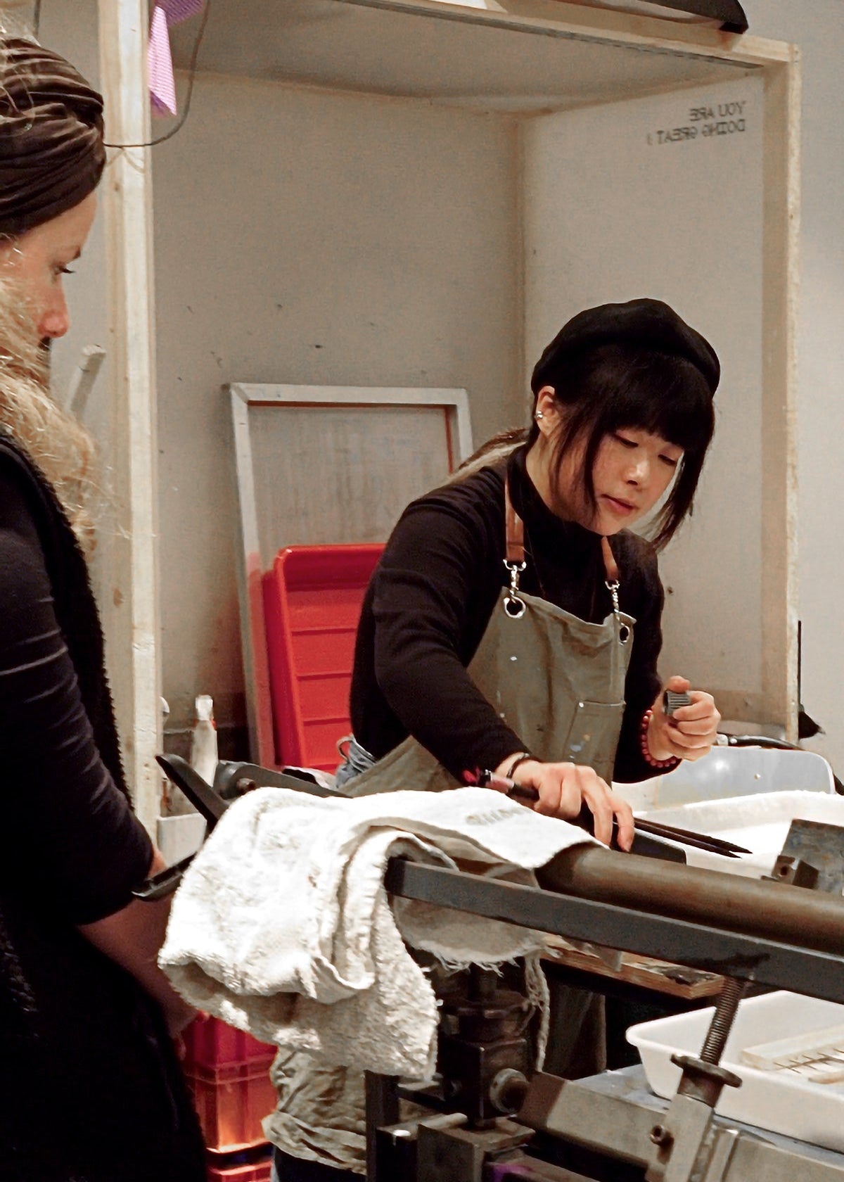 A woman in a black beret and gray apron works at a printing press inside a studio, with another person partially visible on the left.