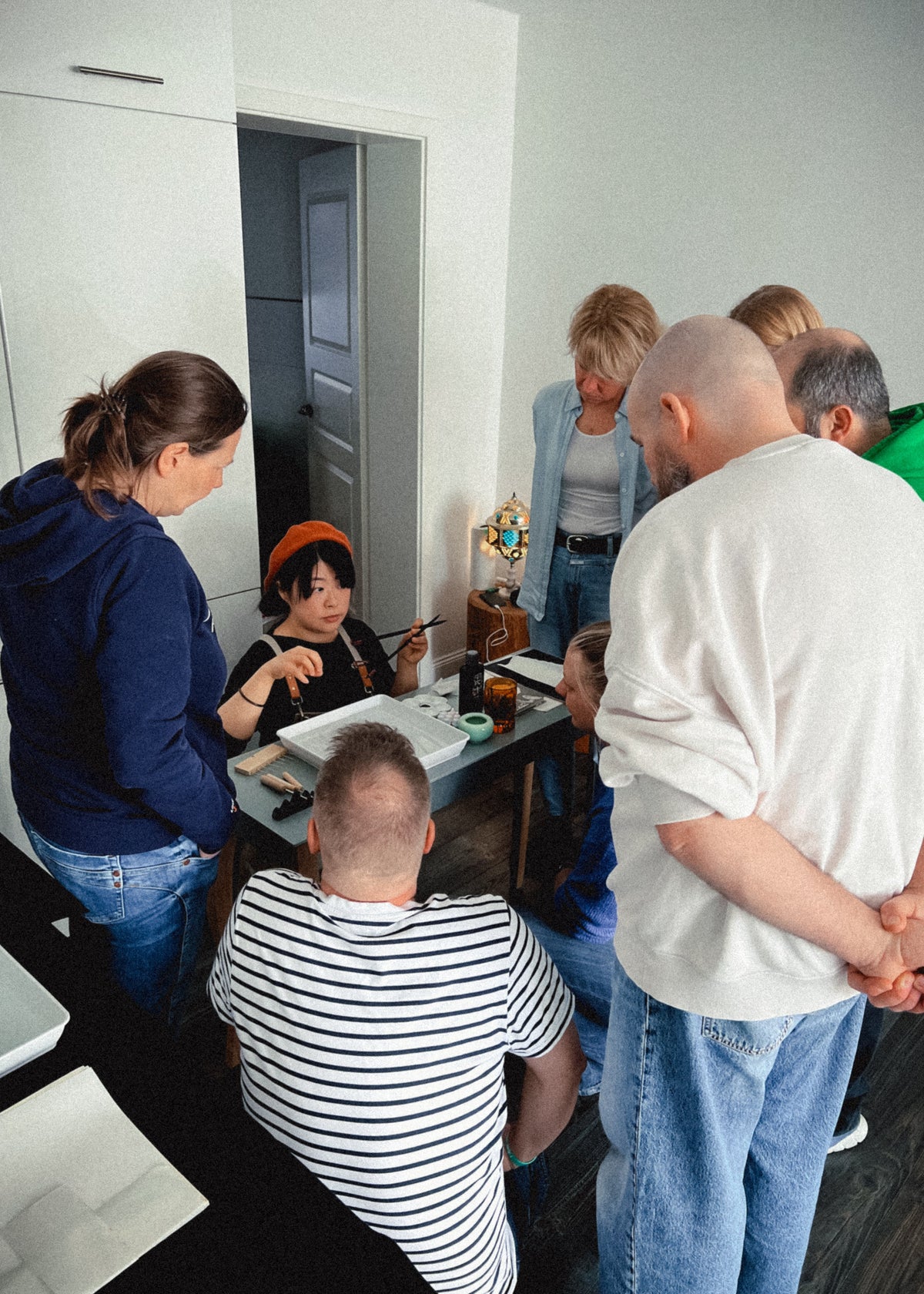 A group of people gathered around a table in a modern indoor setting, with one person seated at the table and several others standing and leaning in closely. The room has light-colored walls and a dark floor.