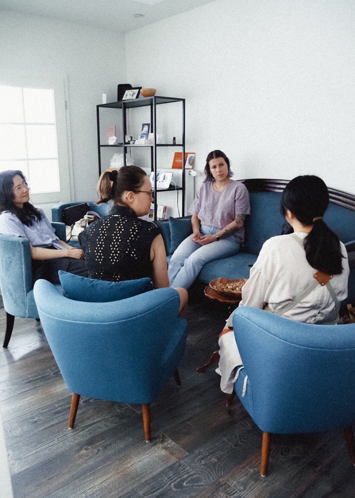 Four people seated in a bright, modern room with blue armchairs and a dark wood floor, engaged in conversation. The room features white walls, a black shelving unit, and a window on the left.