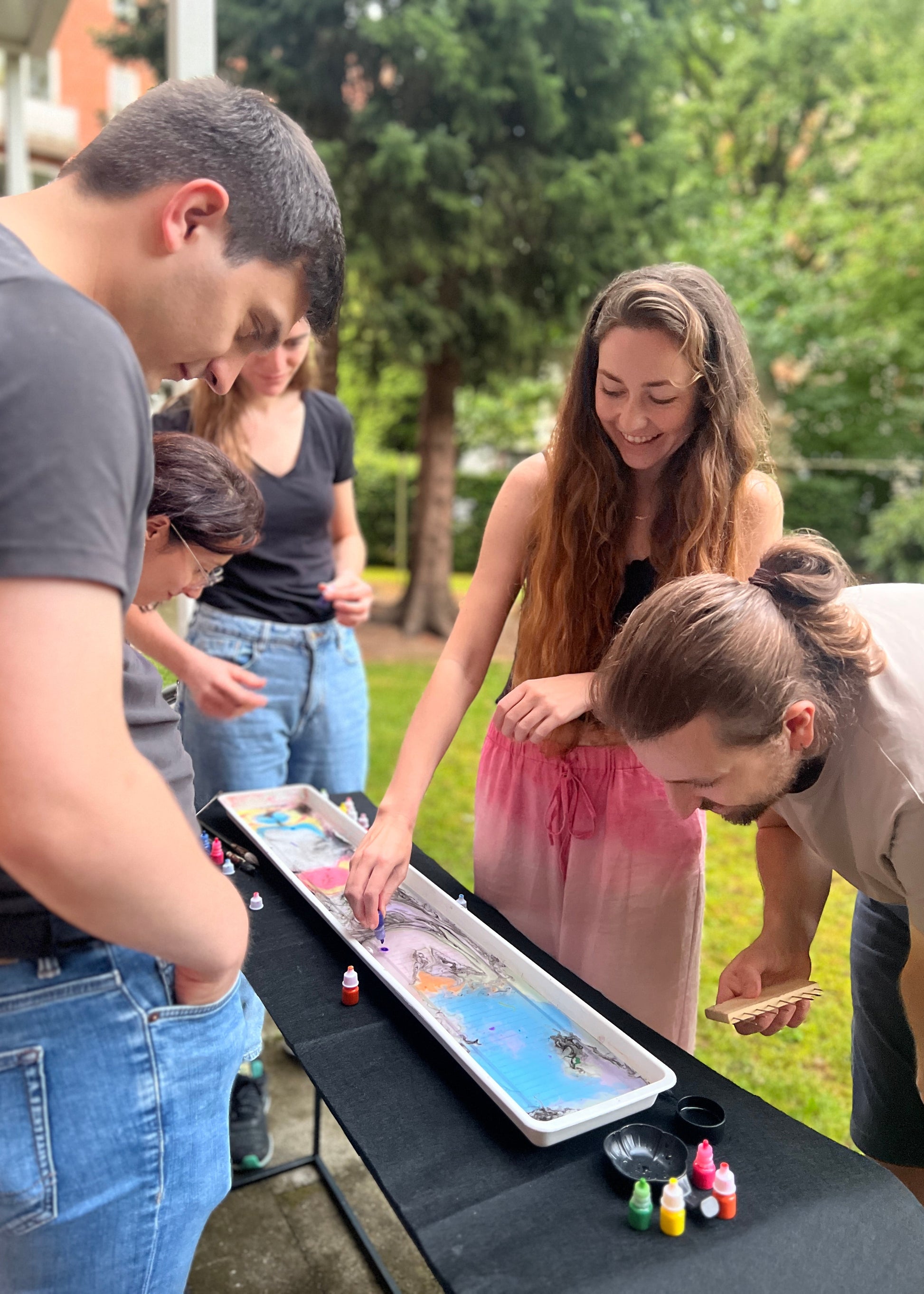 A group of four people gathered outdoors around a long tray filled with colorful paint or ink, painting or writing on it. The scene is set in a grassy outdoor area with trees in the background.