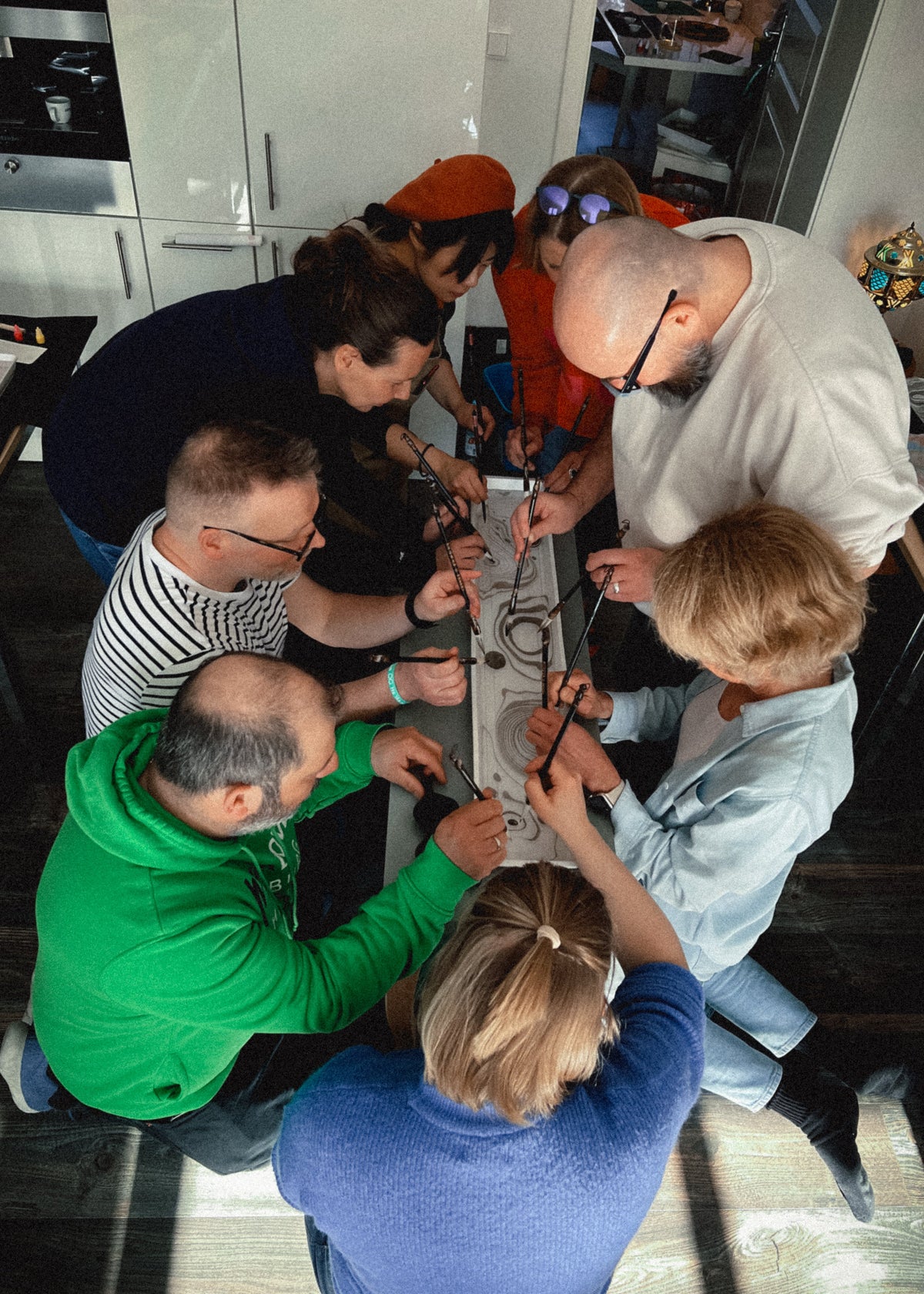 A group of seven people gathered closely around a long, narrow white object laid on the floor in a modern indoor room. The scene is shot from above, showing the group leaning in to examine the item, which appears to be a long, thin white device with black components and cords.
