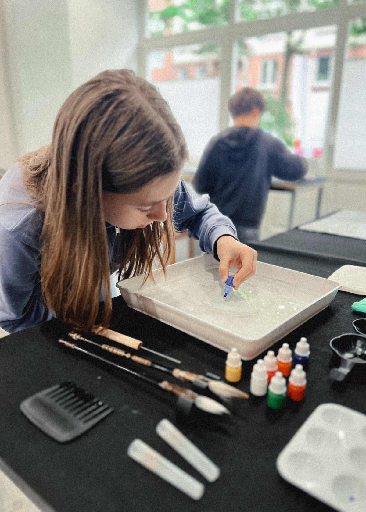 A woman leans over a table with water, mixing something in a tray while paint brushes, small bottles of color, and a palette sit on the table. Another person is blurred in the background near large windows.