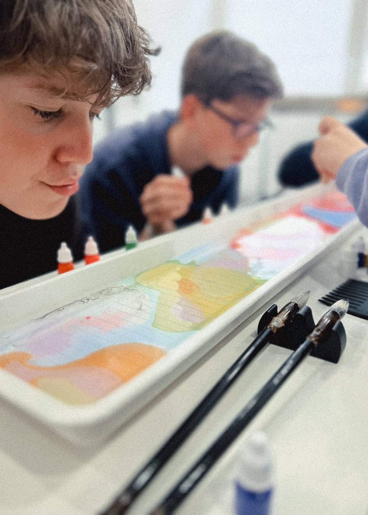 Students painting in a classroom setting, with one boy in the foreground looking down at a long white tray filled with colorful paint and brushes resting on the table.