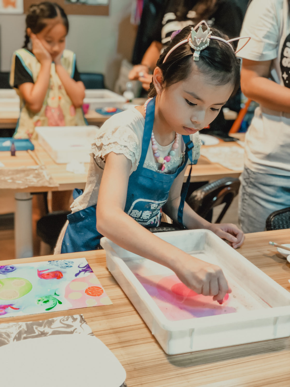 A young girl wearing a blue apron and a tiara sits at a wooden table, dipping a piece of fabric into a tray of colorful water. Other children and adults are visible in the background of what appears to be a craft or art class.