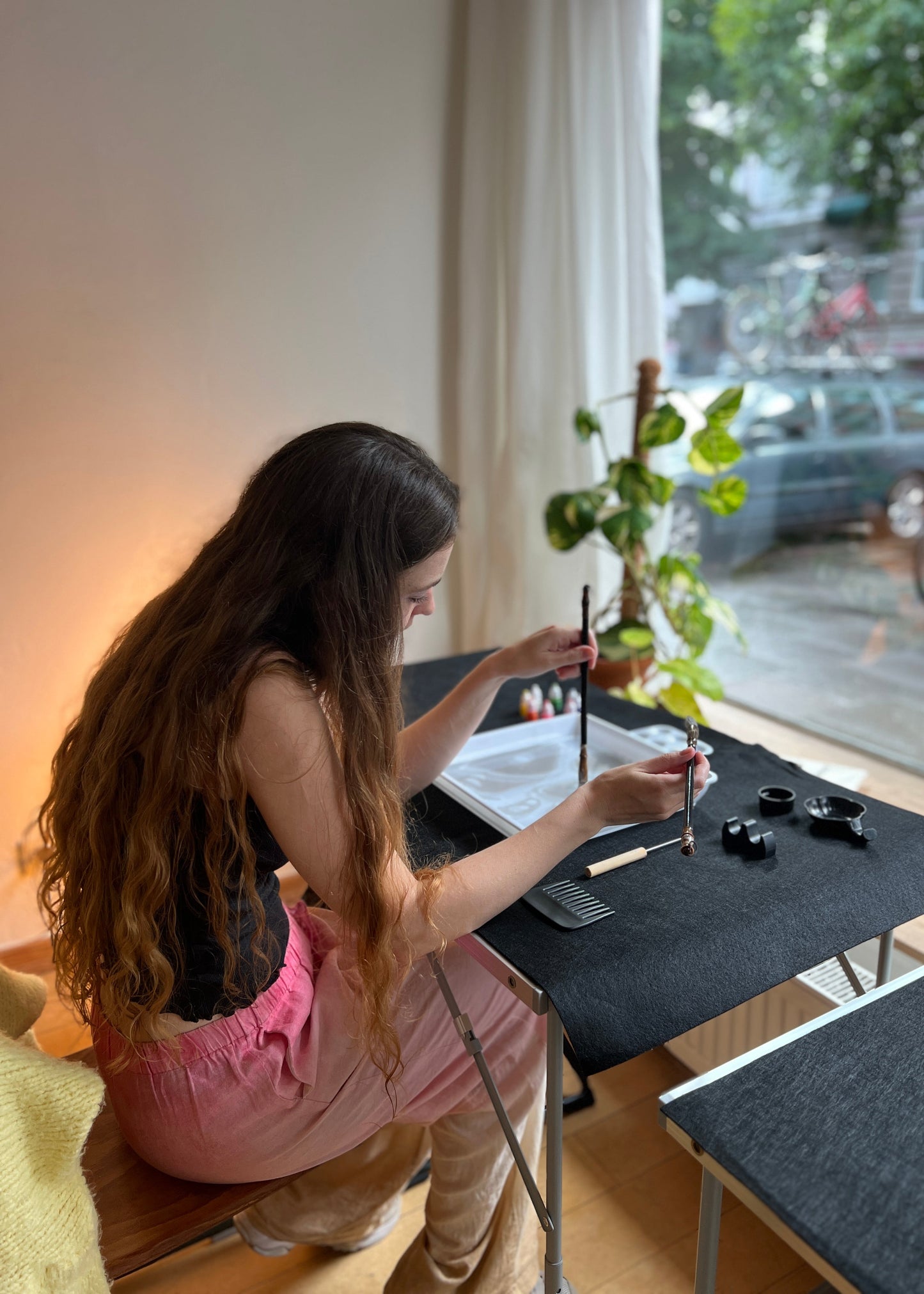 A woman with long wavy brown hair sits at a small table by a window, writing or painting with a brush. The table has ink containers and paper, and a potted plant stands nearby.