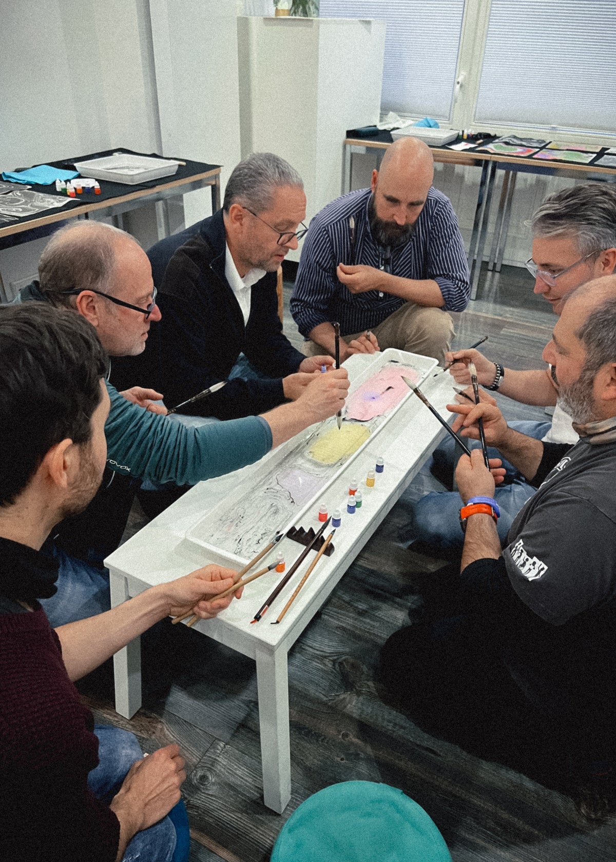 A group of men gathered around a white table in an indoor studio or workshop, painting on a long canvas with brushes and small paint tubes.