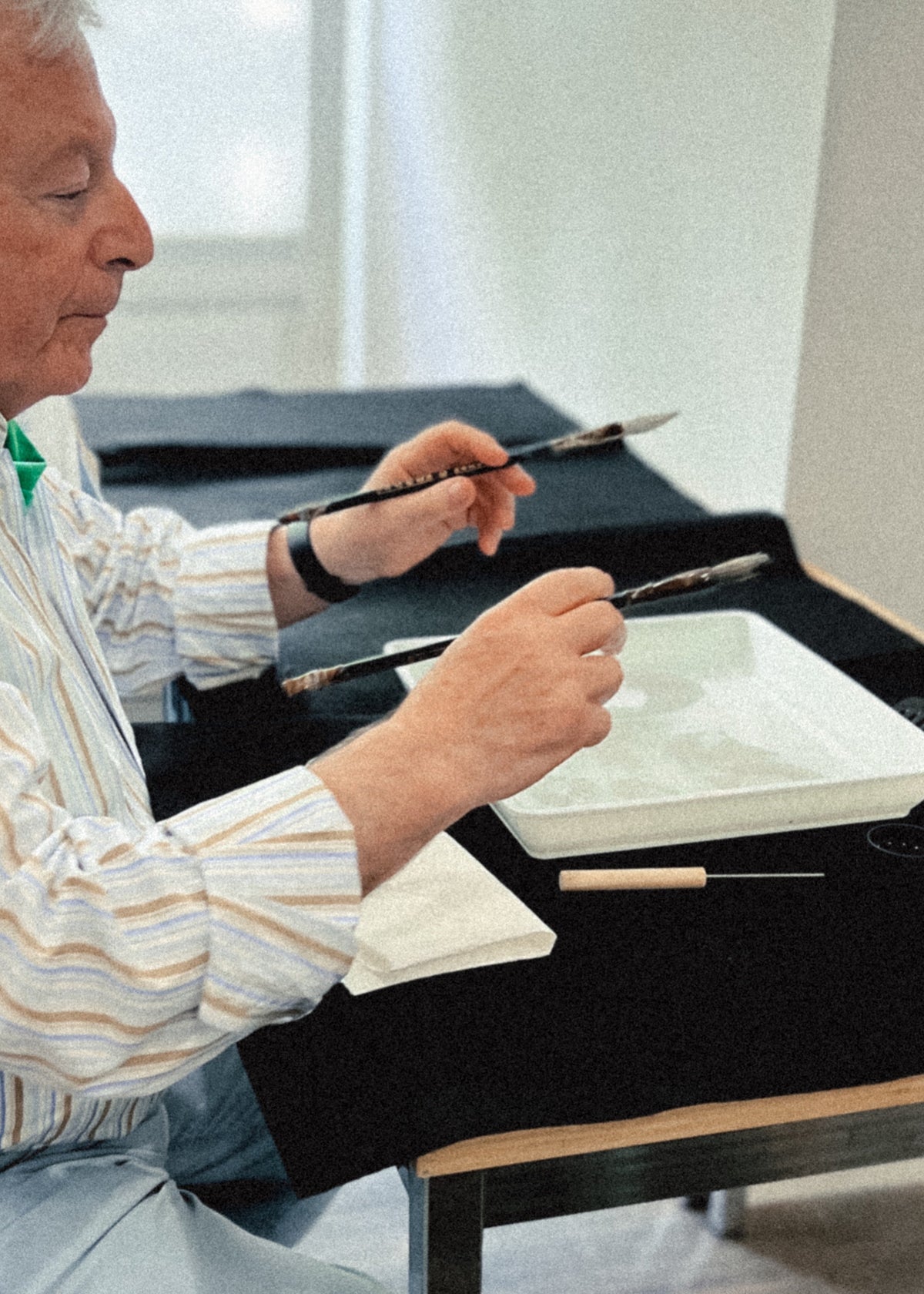 An older man seated at a table, holding two calligraphy brushes over a white ink tray. He wears a striped long-sleeve shirt, and the scene is photographed in a softly lit indoor setting.