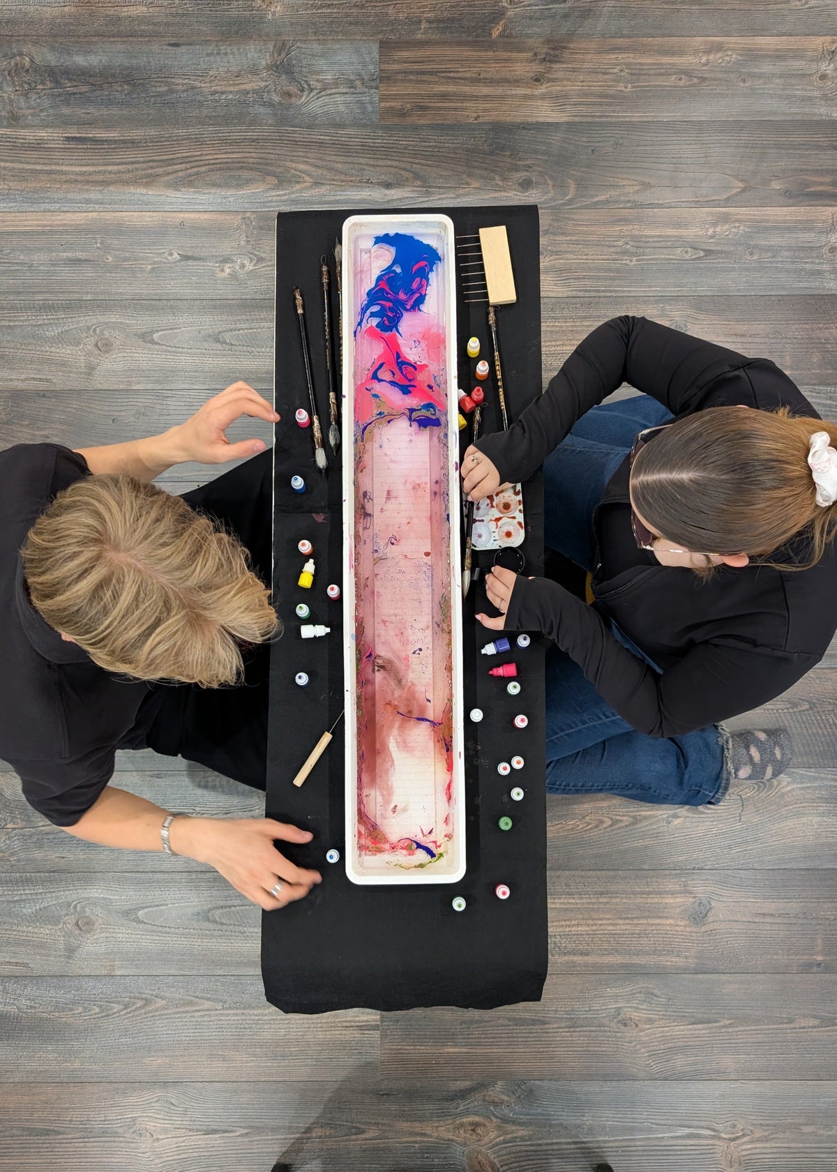 Overhead view of two people kneeling around a long rectangular table on a wood floor, working with a large marbled pink, blue, and white paint tray and various paint tubes and brushes.