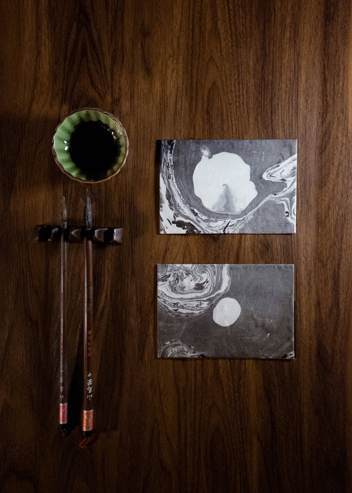 Two rectangular black-and-white abstract art prints with circular white forms, displayed on a dark wood surface next to a small green bowl and two hanging calligraphy brushes.