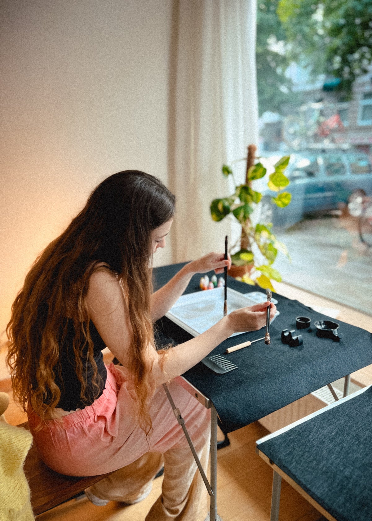 A woman with long dark hair sits at a small table by a window, using a traditional brush to write or paint on paper. She wears a pink top and light pants, with ink pots and a plant nearby in a softly lit indoor setting.